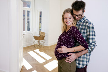 Young man embracing pregnant woman in new home with pram in background - CHAF000584
