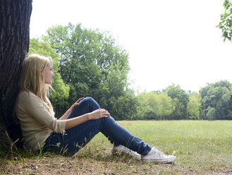 Junge Frau sitzt auf einer Wiese und lehnt sich an einen Baumstamm - BFRF001328