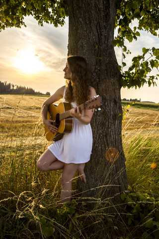 Junge Frau mit Gitarre an Baumstamm gelehnt, Gerstenfeld am Abend, lizenzfreies Stockfoto