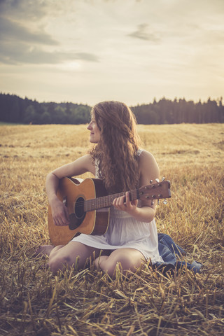 Junge Frau mit Gitarre auf einem Gerstenfeld am Abend sitzend, lizenzfreies Stockfoto
