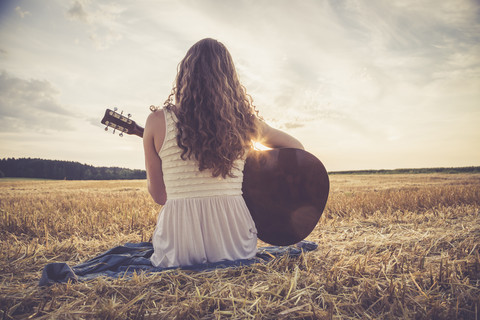 Junge Frau mit Gitarre auf einem Gerstenfeld am Abend sitzend, lizenzfreies Stockfoto