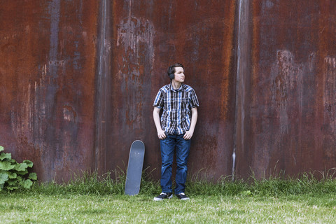 Teenage boy with skateboard leaning on corten wall hearing music stock photo