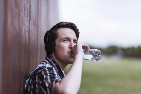 Jugendlicher mit Kopfhörern, der sich an eine Cortenwand lehnt und Wasser trinkt - MMFF000862