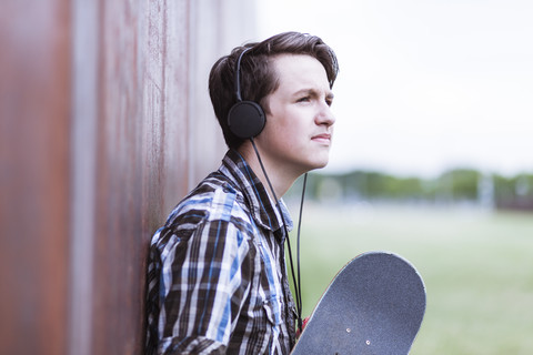 Jugendlicher mit Skateboard, der sich an eine Cortenwand lehnt und Musik hört, lizenzfreies Stockfoto