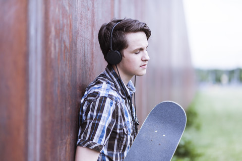 Jugendlicher mit Skateboard, der sich an eine Cortenwand lehnt und Musik hört, lizenzfreies Stockfoto
