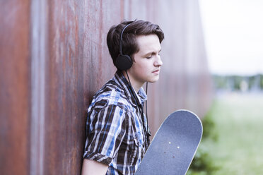 Jugendlicher mit Skateboard, der sich an eine Cortenwand lehnt und Musik hört - MMFF000864