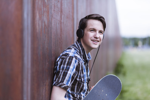 Teenage boy with skateboard leaning on corten wall hearing music stock photo