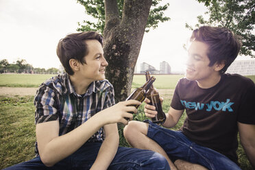 Germany, Berlin, two teenage boys sitting under a tree toasting with beer bottles - MMFF000879