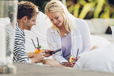 Junges Paar sitzt in einem Café im Freien mit Gläsern mit frischem Saft und schaut auf sein Smartphone - CHAF000956