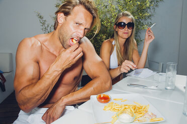 Paar sitzt im Sommer am Tisch, isst Pommes frites und raucht eine Zigarette - CHAF000623
