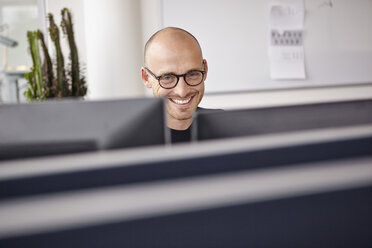 Lächelnder Mann im Büro hinter Computerbildschirmen - RHF000954