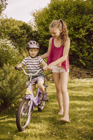 Mädchen hilft kleinem Jungen beim Fahrradfahren im Garten, lizenzfreies Stockfoto
