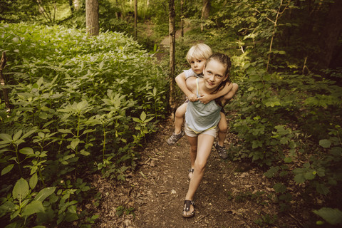 Mädchen trägt kleinen Jungen huckepack durch einen Wald, lizenzfreies Stockfoto