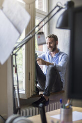 Lächelnder junger Mann im Büro, der am Fenster sitzt - FKF001342