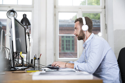 Junger Mann im Büro mit Kopfhörern bei der Arbeit am Computer - FKF001234