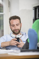 Junger Mann im Büro mit Füßen auf dem Schreibtisch, der auf sein Handy schaut - FKF001237