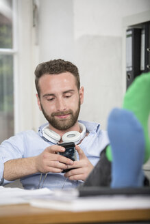 Junger Mann im Büro mit Füßen auf dem Schreibtisch, der auf sein Handy schaut - FKF001237