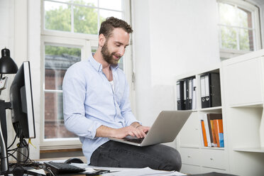 Lächelnder junger Mann im Büro, der am Schreibtisch sitzt und einen Laptop benutzt - FKF001238