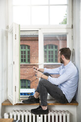 Junger Mann mit Handy sitzt am Fenster und schaut hinaus - FKF001324