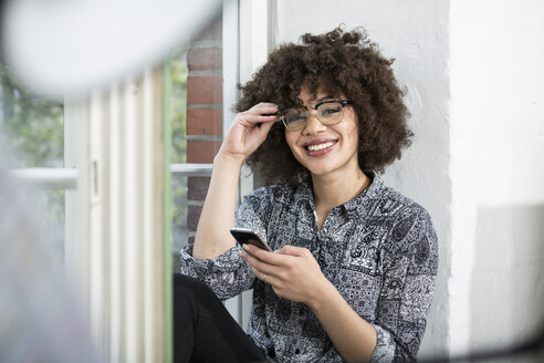 Portrait of smiling young woman with cell phone at the window - FKF001242