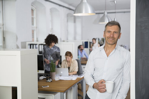 Smiling man in office with colleagues in background - FKF001270