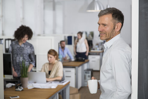 Smiling man in office with colleagues in background - FKF001272