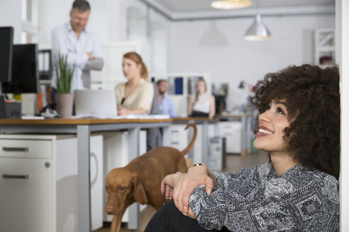 Smiling woman in office with colleagues and dog in background - FKF001329