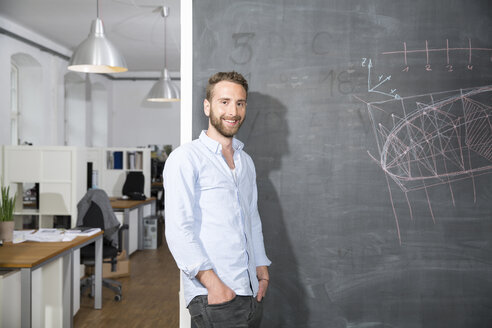 Smiling young man standing at blackboard in office - FKF001287