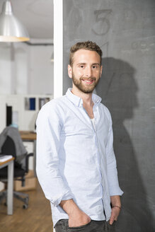 Smiling young man standing at blackboard in office - FKF001288