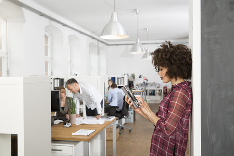 Frau im Büro mit digitalem Tablet und Kollegen im Hintergrund, lizenzfreies Stockfoto