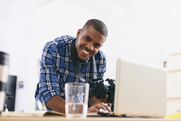 Porträt eines lächelnden jungen Mannes bei der Arbeit mit einem Laptop in seinem Fotostudio - EBSF000796