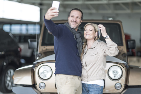 Glückliches Paar im Autohaus, das ein Selfie vor seinem neuen Auto macht, lizenzfreies Stockfoto