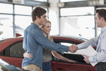 Car dealer and young couple shaking hands in showroom - ZEF006933