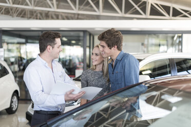 Car dealer showing brochure to young couple in showroom - ZEF006940