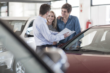 Car dealer showing brochure to young couple in showroom - ZEF006945