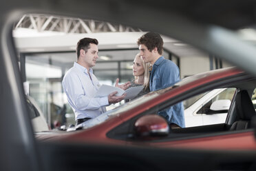 Autohändler zeigt einem jungen Paar im Ausstellungsraum eine Broschüre - ZEF006946