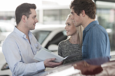 Car dealer showing brochure to young couple in showroom - ZEF006947