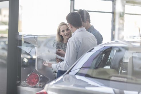 Autohändler zeigt einem jungen Paar im Ausstellungsraum eine Broschüre - ZEF006955