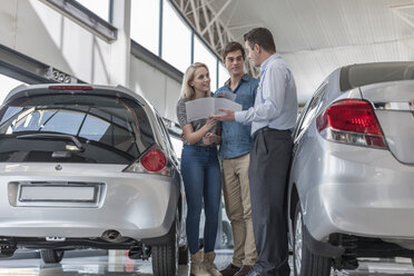 Car dealer showing brochure to young couple in showroom - ZEF007015