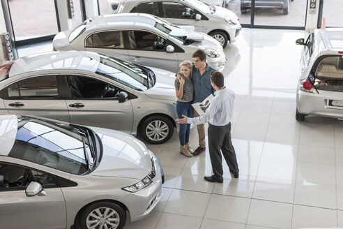 Car dealer showing new car to young couple in showroom - ZEF006965