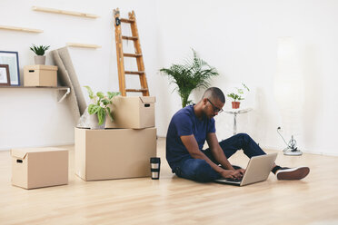 Young man sitting beside cardboard boxes in his new flat using laptop - EBSF000808