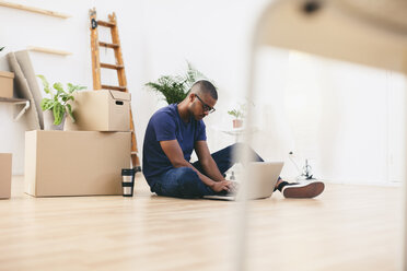 Junger Mann sitzt neben Kartons in seiner neuen Wohnung und benutzt einen Laptop - EBSF000809