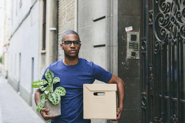 Spain, Barcelona, portrait of young man with cardboard box and foliage plant in front of a house - EBSF000817