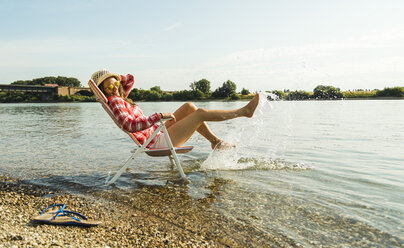 Junge Frau sitzt auf einem Liegestuhl im Fluss und spritzt mit Wasser - UUF005025