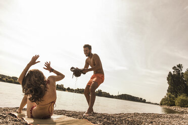 Verspielter junger Mann, der versucht, Wasser über seine Freundin zu gießen, die am Flussufer ein Sonnenbad nimmt - UUF005036