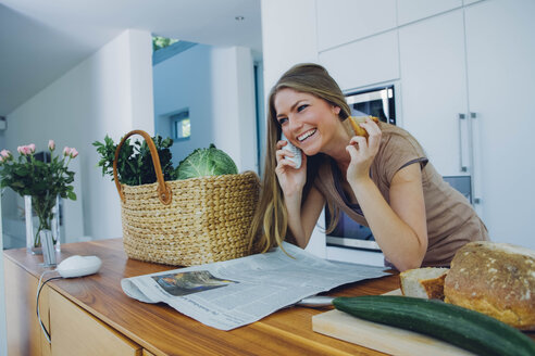 Happy woman on phone in kitchen taking groceries from bag - CHAF000838