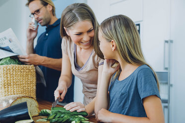 Mother and daughter in kitchen slicing cucumber with father in background - CHAF000844