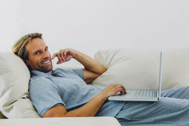 Smiling man lying on couch using laptop - CHAF000975