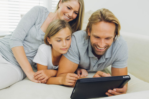 Glücklicher Vater, Mutter und Tochter liegen auf der Couch und teilen sich ein digitales Tablet, lizenzfreies Stockfoto