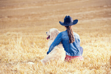 Back view of young woman and Golden Retriever sitting on a stubble field - MAEF010825